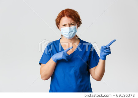 Medical workers, covid-19 pandemic, coronavirus concept. Skeptical and unamused redhead female doctor, physician looking with judgement, pointing upper right corner with reluctant face, wear mask 69156378