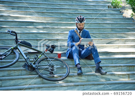 Businessman with bicycle sitting on stairs and using digital tab Businessman with bicycle sitting on stairs and using digital tab 69156720
