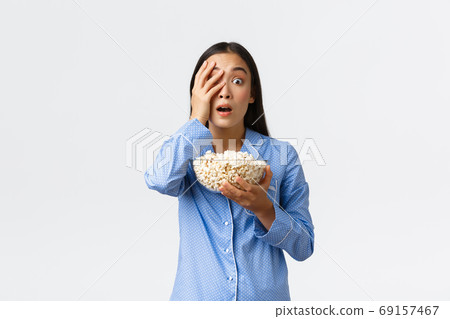 Home leisure, sleepover and slumber party concept. Shocked asian girl in pajamas holding popcorn, gasping and looking startled, open mouth with interest as watching tv or movie, white background 69157467