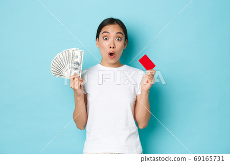 Shopping, money and finance concept. Amused asian girl in white casual t-shirt gasping, found out awesome prices, discount offers in shop, holding both credit card and cash, light-blue background Shopping, money and finance concept. Amused asian girl in white casual t-shirt gasping, found out awesome prices, discount offers in shop, holding both credit card and cash, light-blue background 69165731