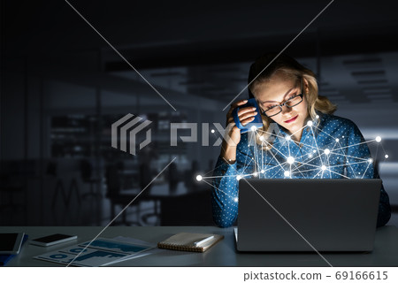 Attractive blonde wearing glasses in dark office using laptop. Mixed media Attractive blonde wearing glasses in dark office using laptop. Mixed media 69166615