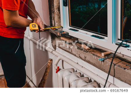 The man performs installation work with the window, for this he also uses a tape measure, measures distances and fastens the window to the wall. 69167649