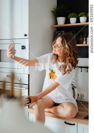 Young pretty woman drinks coffee sitting at her kitchen at home and taking selfie. Smiling woman taking selfie. Young pretty woman drinks coffee sitting at her kitchen at home and taking selfie. Smiling woman taking selfie. 69169993