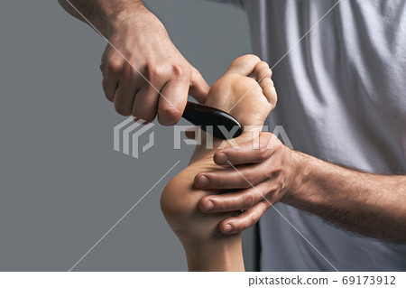 Male masseur hands doing massage on female foot reflex zone in the spa salon using gua sha scraping acupuncture tool. Male masseur hands doing massage on female foot reflex zone in the spa salon using gua sha scraping acupuncture tool. 69173912