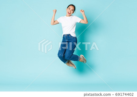 Fashion, beauty and lifestyle concept. Cheerful triumphing, attractive asian girl jumping from happiness and joy, winning competition, celebrating victory over light blue background 69174402