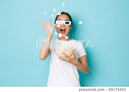 Lifestyle, leisure and emotions concept. Impressed and excited asian girl enjoying watching movie, throwing popcorn at tv screen as wearing 3d glasses, standing light blue background 69174403