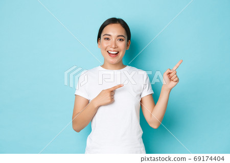 Beautiful happy asian girl in white t-shirt helping with choice, showing way or demonstrating banner. Korean woman looking enthusiastic while pointing upper right corner over blue background 69174404