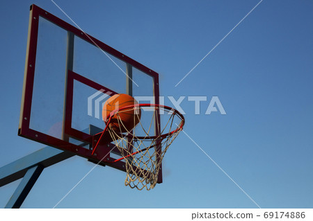 An orange basketball is flying into the basket on an open area against the sky. 69174886