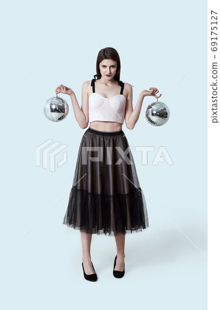 Young beautiful brunette girl in dark box pleated midi skirt posing in studio. Woman balancing holding two disco balls reflecting light. 69175127