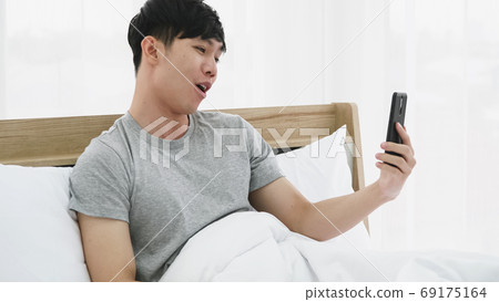 Young happy man using mobile phone for video call in bed at home 69175164