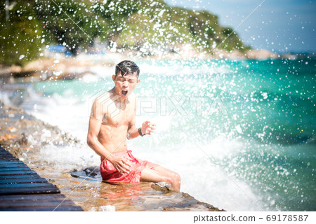 Slim young Asian man sits on a sea pier with the excited face & expressive hand gesture being wet under shower of splash after a large wave hit at a sunny day on Koh Samet, Thailand 69178587