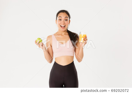 Sport, wellbeing and active lifestyle concept. Excited cute asian fitness girl, sportswoman with apple and orange juice gasping amazed and happy, eating healthy to stay fit, white background 69202072