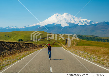 A woman walks along the road with her arms outstretched, panorama against the background of mount Elbrus, Caucasus Russia. A woman walks along the road with her arms outstretched, panorama against the background of mount Elbrus, Caucasus Russia. 69204779
