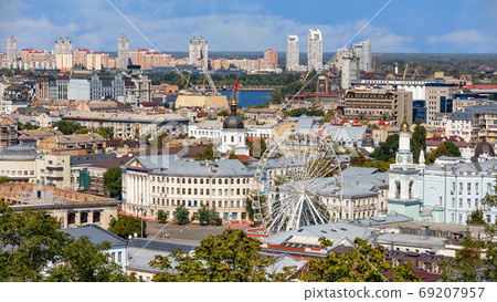 The landscape of the summer city of Kyiv overlooking the old district of Podil with a Ferris wheel and a bell tower with a gilded dome, the Dnipro River and many city buildings. The landscape of the summer city of Kyiv overlooking the old district of Podil with a Ferris wheel and a bell tower with a gilded dome, the Dnipro River and many city buildings. 69207957
