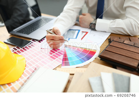 Man sits at table holding pen in his hands on table lie smartphone laptop color palette and a yellow hard hat. Man sits at table holding pen in his hands on table lie smartphone laptop color palette and a yellow hard hat. 69208746
