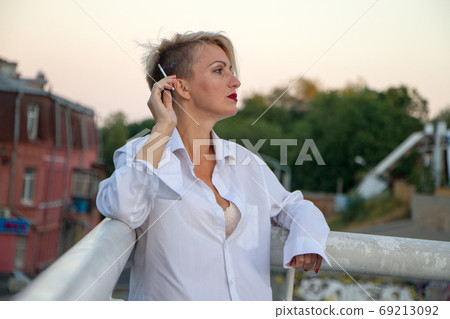 Beautiful mature blonde woman in white shirt smokes white cigarette outdoors in city at summer Beautiful mature blonde woman in white shirt smokes white cigarette outdoors in city at summer 69213092