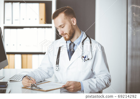 Friendly red-bearded doctor sitting and writing at clipboard in clinic. Perfect medical service in hospital. Medicine concept Friendly red-bearded doctor sitting and writing at clipboard in clinic. Perfect medical service in hospital. Medicine concept 69214292