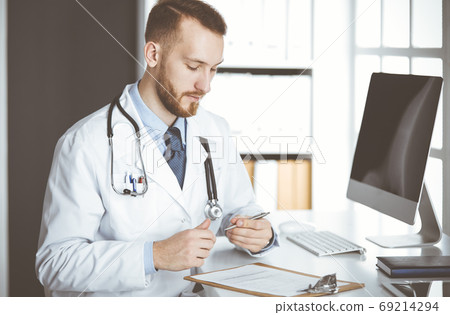 Friendly red-bearded doctor sitting and writing at clipboard in clinic. Perfect medical service in hospital. Medicine concept Friendly red-bearded doctor sitting and writing at clipboard in clinic. Perfect medical service in hospital. Medicine concept 69214294
