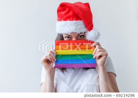 Beautiful lesbian girl in red Santa Claus hat with LGBT rainbow flag isolated on white background looking happy and excited. Young woman Gay Pride portrait. Happy Christmas and New Year holidays Beautiful lesbian girl in red Santa Claus hat with LGBT rainbow flag isolated on white background looking happy and excited. Young woman Gay Pride portrait. Happy Christmas and New Year holidays 69214296