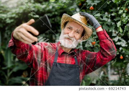 Pleasant positive joyful elderly man gardener in straw hat, posing in beautiful greenhouse with exotic plants and palm trees while making selfie photo or showing video using his smartphone. Pleasant positive joyful elderly man gardener in straw hat, posing in beautiful greenhouse with exotic plants and palm trees while making selfie photo or showing video using his smartphone. 69214743