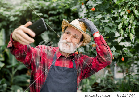 Pleasant positive joyful elderly man gardener in straw hat, posing in beautiful greenhouse with exotic plants and palm trees while making selfie photo or showing video using his smartphone. Pleasant positive joyful elderly man gardener in straw hat, posing in beautiful greenhouse with exotic plants and palm trees while making selfie photo or showing video using his smartphone. 69214744