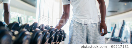 African American man picking up a dumbbell from the rack and looking outside at gym 69217181