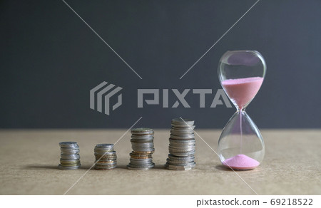 Hourglass with sand flowing and stacked coins on table. Hourglass with sand flowing and stacked coins on table. 69218522