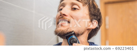 Handsome young bearded man trimming his beard with a trimmer BANNER, LONG FORMAT Handsome young bearded man trimming his beard with a trimmer BANNER, LONG FORMAT 69223401