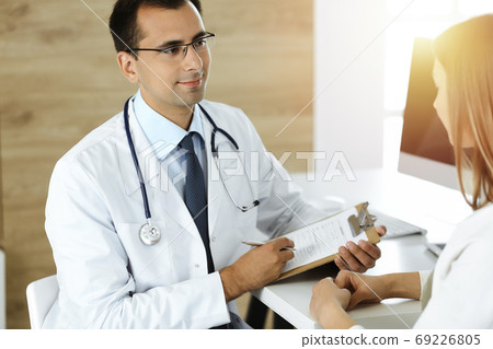 Doctor and patient discussing medical exam results while sitting at the desk in sunny clinic Doctor and patient discussing medical exam results while sitting at the desk in sunny clinic 69226805