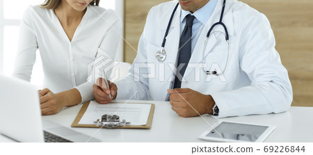 Doctor and patient discussing medical exam results while sitting at the desk in clinic, close-up. Male physician using clipboard for filling up medication history record of young woman 69226844