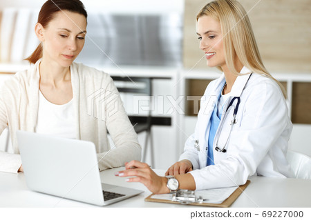 Woman-doctor at work in sunny hospital is happy to consult female patient. Blonde physician checks medical history record and exam results while using clipboard Woman-doctor at work in sunny hospital is happy to consult female patient. Blonde physician checks medical history record and exam results while using clipboard 69227000