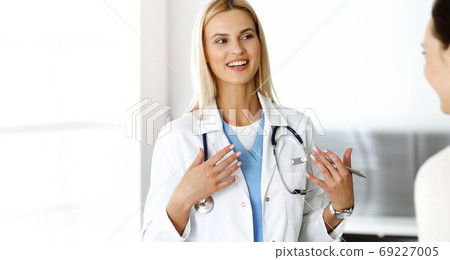 Woman-doctor at work in sunny hospital is happy to consult female patient. Blonde physician checks medical history record and exam results while using clipboard Woman-doctor at work in sunny hospital is happy to consult female patient. Blonde physician checks medical history record and exam results while using clipboard 69227005