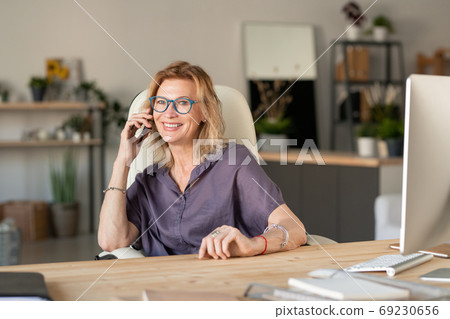 Happy mature businesswoman in casualwear sitting by desk in front of computer 69230656