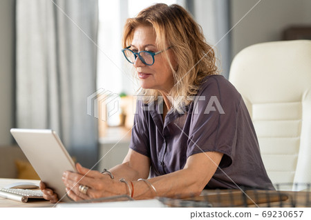 Mature businesswoman in casualwear and eyeglasses looking at tablet display 69230657