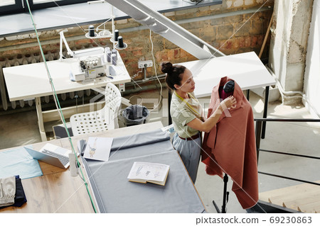 Young fashion designer working with textile on dummy while standing by table 69230863