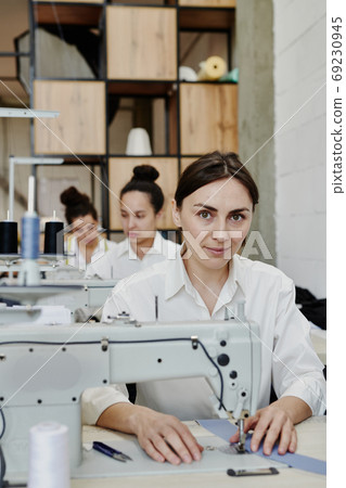 Young pretty seamstress in workwear looking at you while repairing clothes Young pretty seamstress in workwear looking at you while repairing clothes 69230945