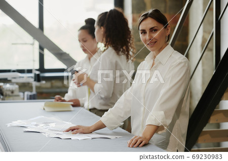 Young creative female designer of clothes looking at you while standing by table 69230983