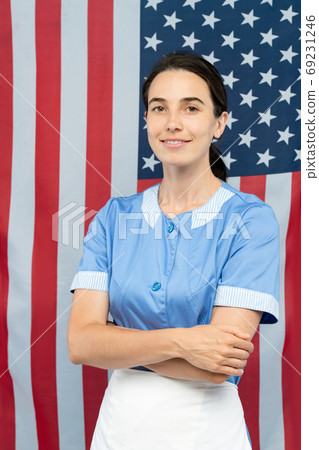 Young cheerful pretty chambermaid in blue uniform crossing arms by chest Young cheerful pretty chambermaid in blue uniform crossing arms by chest 69231246