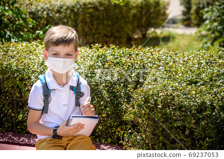 One pupil boy wearing mask is sitting on bench near his school during covid-19 pandemic time. New normal and education concept of coronavirus epidemic. 69237053