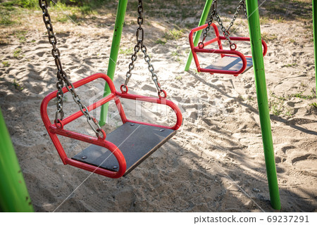 Empty swing-set on playground because of COVID lockdown. Sand underneath the swings and a grassy area, then many trees behind. Good summer camp image 69237291