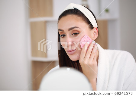 brunette in a bathrobe washes off makeup with a pink sponge brunette in a bathrobe washes off makeup with a pink sponge 69241573