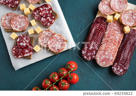 Serving of smoked salchichon and braunschweig sausages in several rows and sandwiches with them are on boards. Next to is cherry tomato and cheese cubes. Flat lay, horizontal is on a green background. Serving of smoked salchichon and braunschweig sausages in several rows and sandwiches with them are on boards. Next to is cherry tomato and cheese cubes. Flat lay, horizontal is on a green background. 69250622