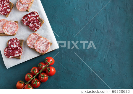 Sandwiches with smoked salchichon and braunschweig sausages are on a paper on the board. Next to are cherry tomatoes on a green background. Flat lay, horizontal composition is on a green background. Sandwiches with smoked salchichon and braunschweig sausages are on a paper on the board. Next to are cherry tomatoes on a green background. Flat lay, horizontal composition is on a green background. 69250623