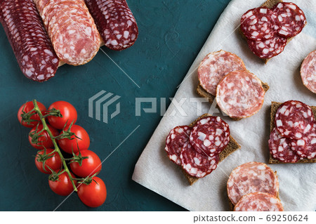 Sandwiches with smoked traditional salchichon and braunschweig sausages are on board. Next to are several rows of sausages and cherry tomatoes on a green background. Flat lay, horizontal composition. Sandwiches with smoked traditional salchichon and braunschweig sausages are on board. Next to are several rows of sausages and cherry tomatoes on a green background. Flat lay, horizontal composition. 69250624