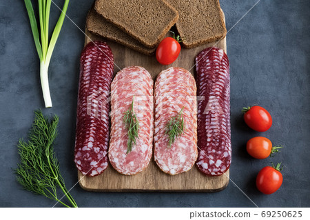 Slices of smoked salchichon and braunschweig sausages lie on a cutting board in several rows. Nearby are cherry tomatoes, green onions, dill and bread. Flat lay on a gray background. Slices of smoked salchichon and braunschweig sausages lie on a cutting board in several rows. Nearby are cherry tomatoes, green onions, dill and bread. Flat lay on a gray background. 69250625