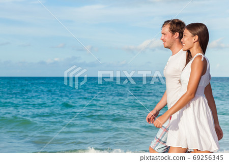 Holiday couple relaxing walking on beach at sunset on honeymoon travel vacation holidays. Young interracial couple in love wearing white dress and shirt, Asian woman and Caucasian man. Holiday couple relaxing walking on beach at sunset on honeymoon travel vacation holidays. Young interracial couple in love wearing white dress and shirt, Asian woman and Caucasian man. 69256345