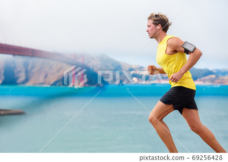 Runner man listening to music during run cardio workout. Running athlete using app on smartphone armband and earphones. Sporty fit young jogger jogging by San Francisco Bay and Golden Gate Bridge. 69256428
