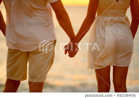 Romantic couple holding hands on beach sunset during travel. Closeup of happy woman and man together in romance on romantic honeymoon getaway in beautiful light. Casual beachwear white linen clothing. Romantic couple holding hands on beach sunset during travel. Closeup of happy woman and man together in romance on romantic honeymoon getaway in beautiful light. Casual beachwear white linen clothing. 69256456