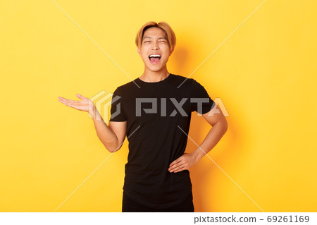 Portrait of happy blond asian guy, winking sassy and smiling, holding something on hand over yellow background Portrait of happy blond asian guy, winking sassy and smiling, holding something on hand over yellow background 69261169