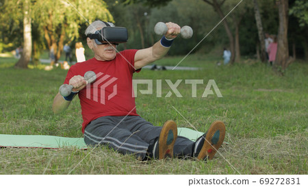Senior old grandfather man in VR headset helmet making fitness exercises with dumbbells in park 69272831